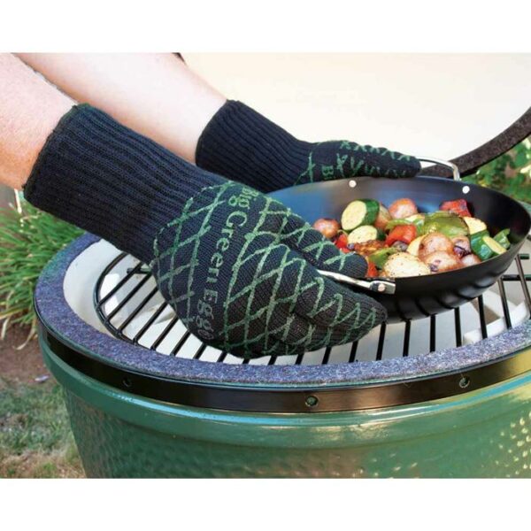 Wearing Pit Mitt BBQ Gloves, two hands hold a grill pan filled with sliced vegetables over an open green barbecue grill outdoors.