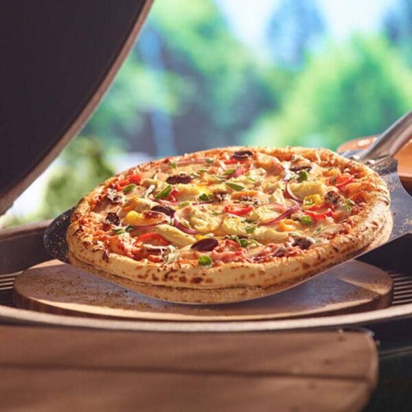 A hot vegetable pizza with peppers, onions, and olives is taken from an outdoor oven with a peel. Its baked on the Pizza/Baking Stone (Large, X-Large & XX-Large) amid a sunlit, green, blurred background.