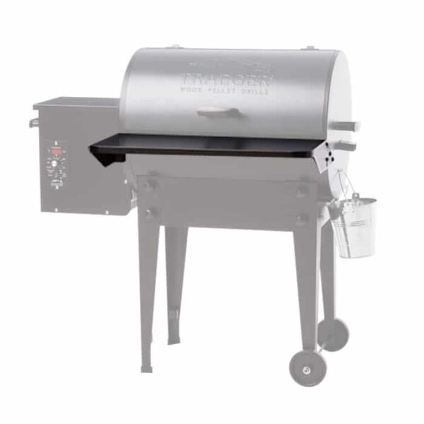 A close-up of the TRAEGER Folding Front Shelf-20 Series attached to a wood pellet grill, with the rest of the grill faded out to highlight the shelf feature. The grill is on wheels and has a small metal bucket hanging on one side.