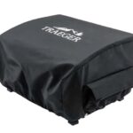 A black Grill Cover - Scout & Ranger is fitted over a grill, featuring the Traeger logo with a white mountain and tree design on top.