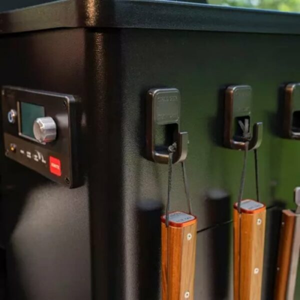 Close-up of Traeger Pink Butcher Paper atop a black outdoor grill, with three wooden-handled barbecue tools hanging from side hooks near the control panel.