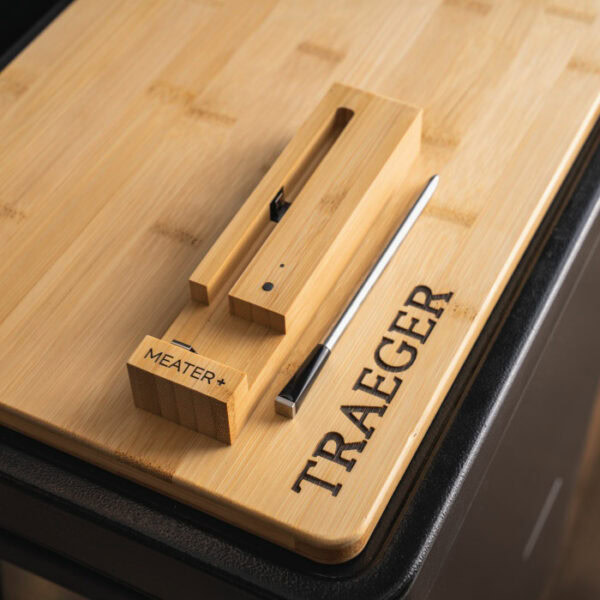 A bamboo cutting board engraved with TRAEGER displays the TRAEGER-Meater+ WiFi Thermometer and its wooden charging case.