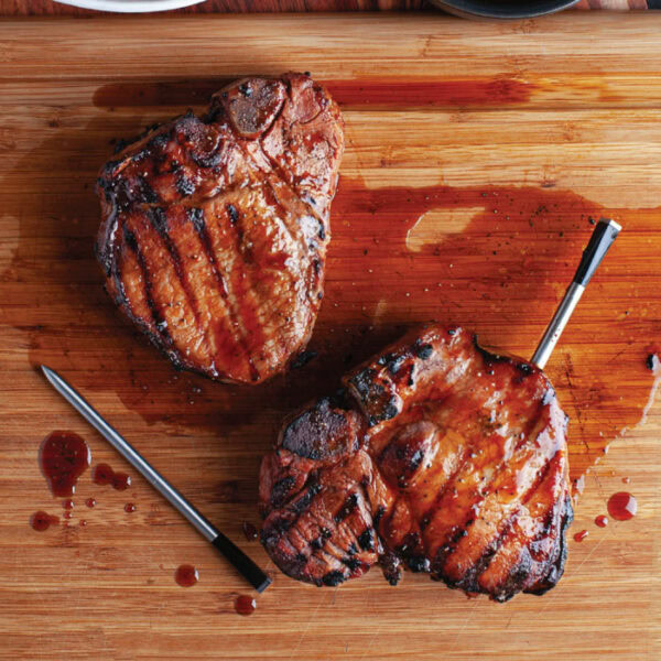 Two grilled steaks with grill marks rest on a wooden cutting board, each with a TRAEGER-Meater+ WiFi Thermometer inserted, and juices pooling around them.