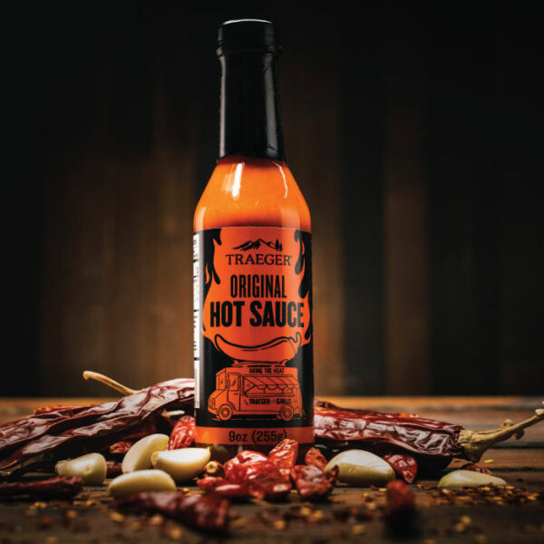 A bottle of Traeger Original Hot Sauce sits on a rustic wooden surface, surrounded by dried red chili peppers and garlic cloves against a dark, blurred background.