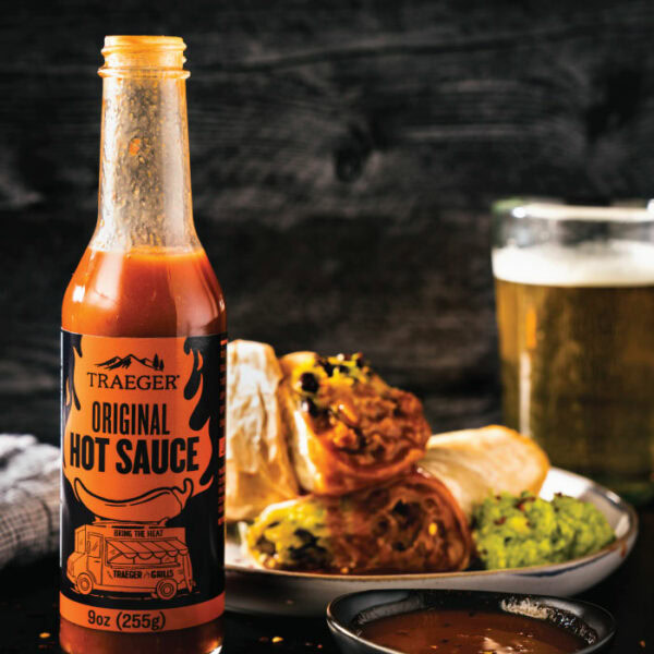 A bottle of Traeger Original Hot Sauce is in front of a plate with burritos, guacamole, and a glass of beer, with a small dish of dipping sauce nearby on a dark rustic background.