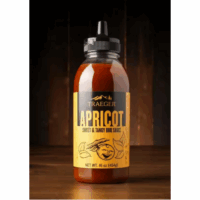 A 16 oz bottle of TRAEGER-Apricot Sweet & Tangy BBQ Sauce, labeled TRAEGER-Apricot 16 OZ Squeeze, with a yellow label sits on a wooden surface against a dark, blurred background.