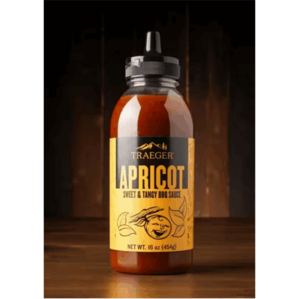 A 16 oz bottle of TRAEGER-Apricot Sweet & Tangy BBQ Sauce, labeled TRAEGER-Apricot 16 OZ Squeeze, with a yellow label sits on a wooden surface against a dark, blurred background.