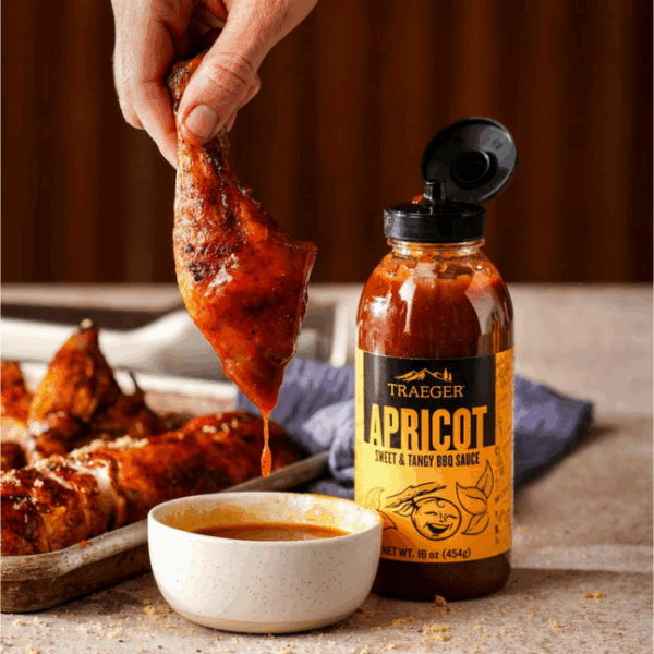 A hand dips grilled chicken into a bowl of sauce beside a bottle of TRAEGER-Apricot 16 OZ Squeeze, with additional chicken pieces on a tray in the background.