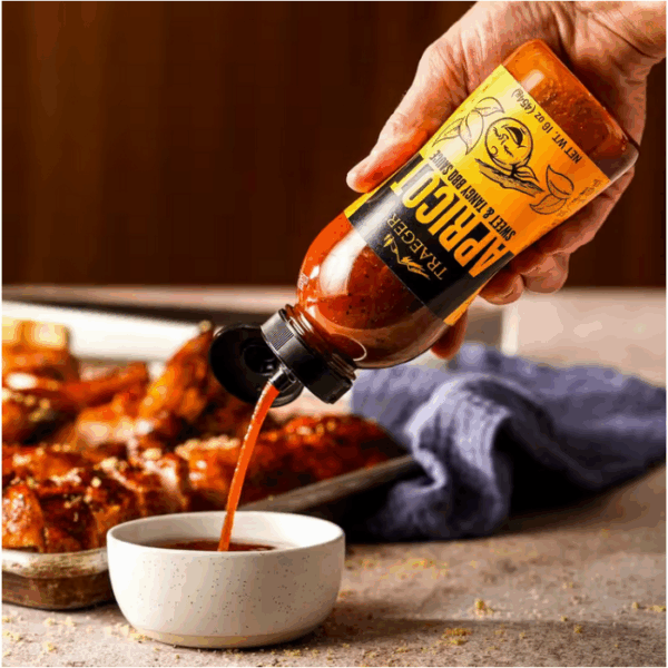 A hand pours TRAEGER-Apricot 16 OZ Squeeze, a bright orange BBQ sauce, into a small white bowl with glazed chicken wings and a baking tray in the background.