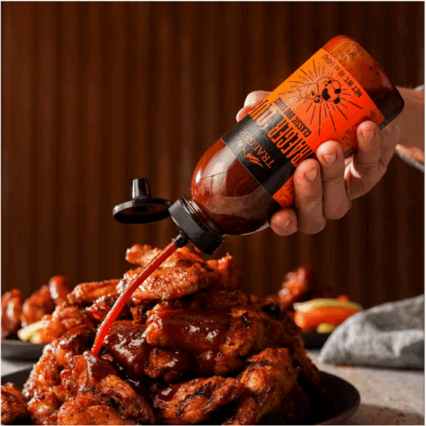 A hand pours Traeger Que Savory BBQ Marinade from a 16 oz. squeeze bottle with an orange label onto saucy chicken wings on a plate, with a blurred brown background and some vegetables in view.