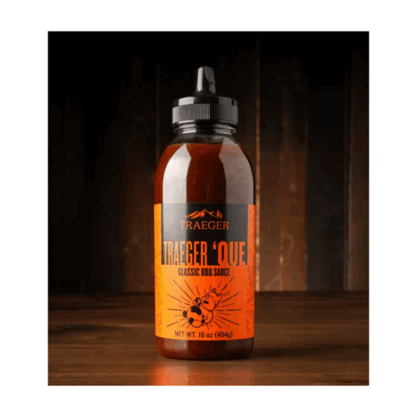 A bottle of Traeger Que Savory BBQ Marinade 16 oz. Squeeze Bottle with an orange label and black flip-top cap is shown on a wooden surface against a dark, blurred background.