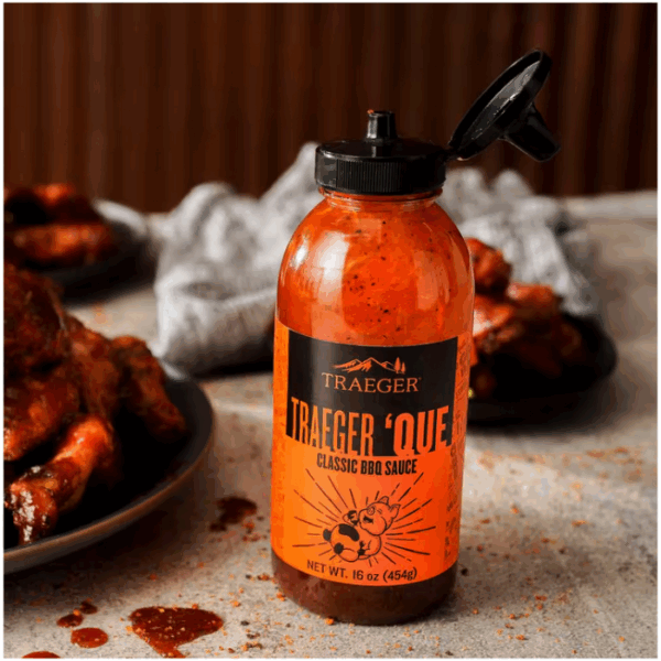 A 16 oz. squeeze bottle of Traeger Que Savory BBQ Marinade sits open on a gray countertop, with sauce spilled nearby and plates of saucy barbecued meat plus a crumpled gray napkin in the background.