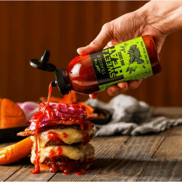 A hand pours TRAEGER-Sweet & Heat 16 OZ Sqz BBQ Sauce onto a cheeseburger stacked with bacon, melted cheese, and red onion on a rustic wooden table, with burger buns and vegetables blurred in the background.
