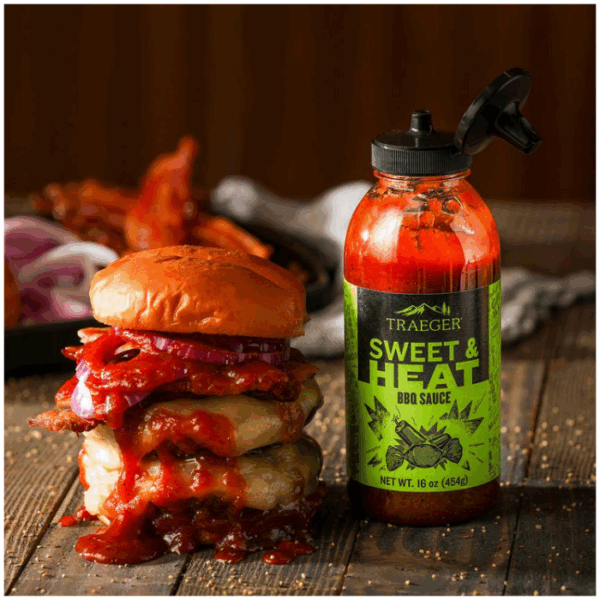 A messy cheeseburger with bacon, onions, and barbecue sauce sits beside a bottle of TRAEGER-Sweet & Heat 16 OZ Sqz on a rustic wooden table.