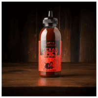 A bottle of TRAEGER-Texas Spicy 16 OZ Sqz BBQ Sauce with a black cap sits on a wooden surface against a dark, rustic wood background. Its red label displays flame graphics and product details.