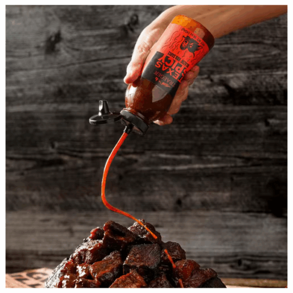 A hand pours TRAEGER-Texas Spicy 16 OZ Sqz barbecue sauce onto saucy, grilled meat chunks, with a rustic dark wooden background.