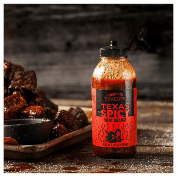 A bottle of TRAEGER-Texas Spicy 16 OZ Sqz rests on a wooden surface beside a tray of grilled meat, set against a rustic, dark wood background.
