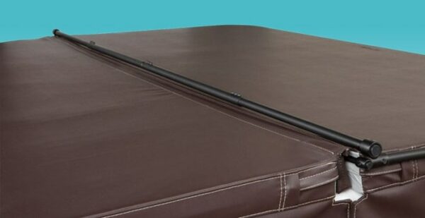A black metal Cover Valet Pro lifter spans across the seam of a closed brown leather hot tub cover with visible stitching and a small tear near the corner, set against a plain light blue background.