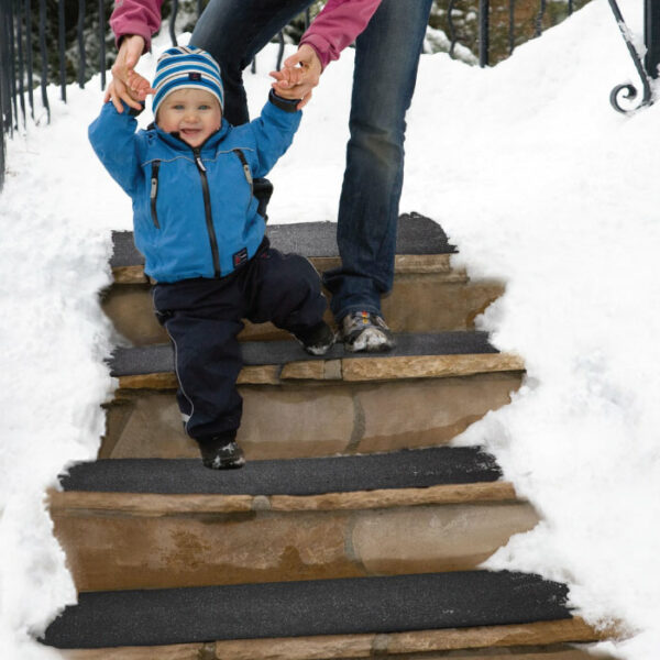 An adult helps a smiling toddler walk down snowy steps covered by a HeatTrak 10 x 30 Heated Stair Mat. The child, in a blue jacket and striped hat, is supported by the adult as snow blankets the ground beside the mat.