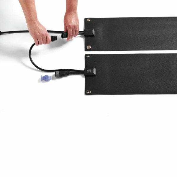 A person’s hands connect a black electrical cord to the HeatTrak 10 x 30 Heated Stair Mat, a flat, rectangular black mat with metal grommets at the corners, set against a white background.