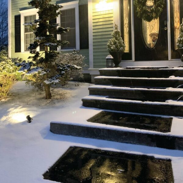 A HeatTrak Heated Snow Melting Entrance Mat (30 x 48) keeps the steps clear of snow, leading to a house with a welcoming wreath and glowing porch lights. A dog looks out the glass door; nearby bushes and tree have a light dusting of snow.