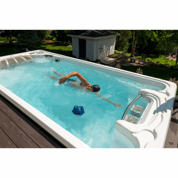 A person enjoys swimming freestyle in an outdoor swim spa with the FROG @EASE FLOATING SYSTEM, surrounded by greenery and a small building on a sunny day.