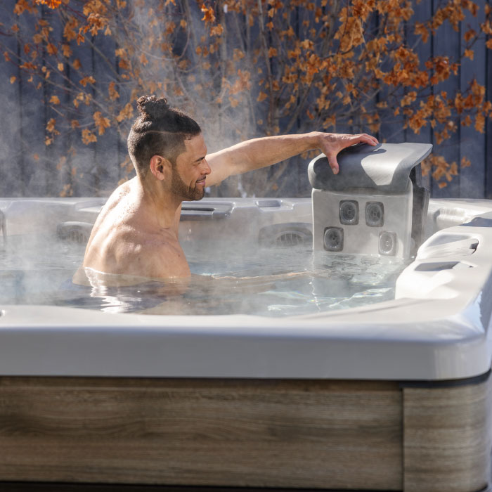 A man unwinds in an A7 PLUS KIT - Platinum Shell / Coastal Grey Cabinet hot tub, adjusting the headrest. Autumn leaves hang nearby while sunlight brightens the steaming outdoor scene.