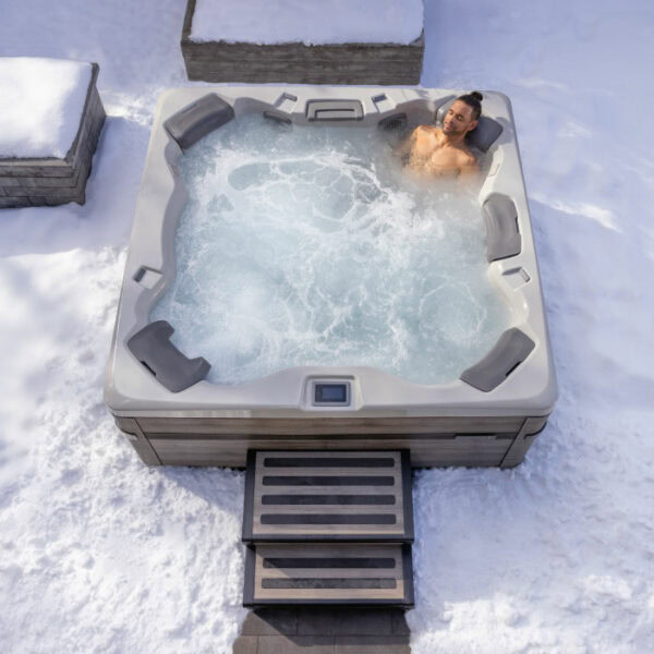 A person relaxes alone in the A7 PLUS KIT - Platinum Shell / Coastal Grey Cabinet hot tub, surrounded by snow with steps leading to the tub and nearby snow-covered benches.