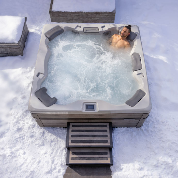 A person relaxes alone in the A7 PLUS KIT - Platinum Shell / Coastal Grey Cabinet hot tub, surrounded by snow with steps leading to the tub and nearby snow-covered benches.