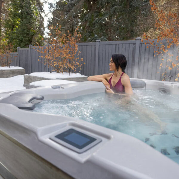 A woman in a maroon swimsuit relaxes in the A8 SELECT KIT hot tub with a Snow Shell and Timber Cabinet, enjoying the steaming water outdoors in winter, surrounded by snow, bare trees with orange leaves, and a gray fence.