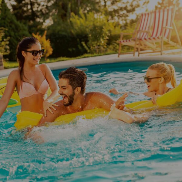 Three young adults in swimsuits and sunglasses laugh and play on yellow floats in a sunlit backyard, enjoying the Above Ground Gold Pool Opening with water splashing and deck chairs visible nearby.
