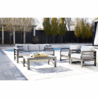 The Amora Grouping 4-piece set, featuring a gray cushioned sofa, two chairs, and a coffee table, is arranged on a rug by the pool amid trees and a black fence on a sunny day.