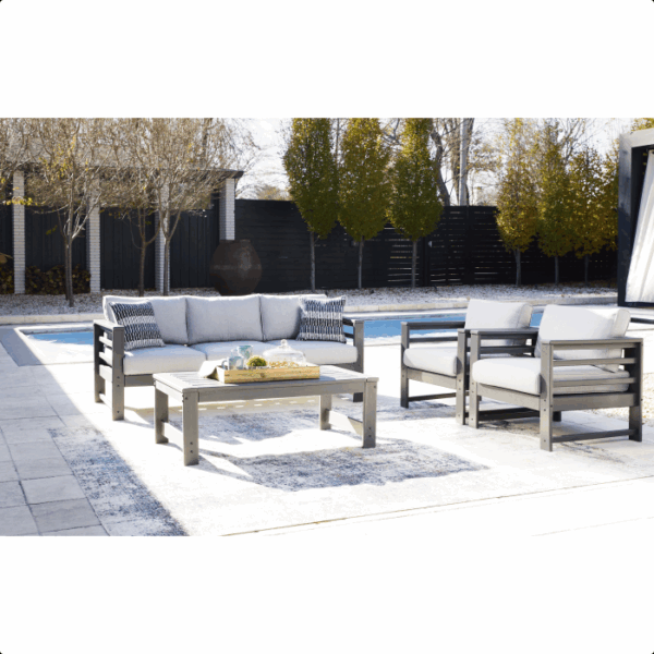 The Amora Grouping 4-piece set, featuring a gray cushioned sofa, two chairs, and a coffee table, is arranged on a rug by the pool amid trees and a black fence on a sunny day.