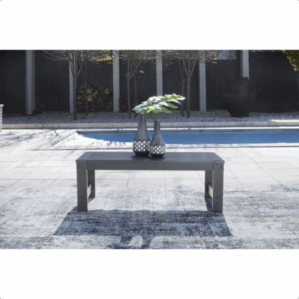 The Amora Grouping 4-piece features a modern outdoor coffee table adorned with two decorative vases and green leaves, set on a patterned rug overlooking a swimming pool and minimalist garden.