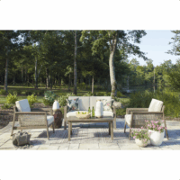 The Barn Cove Grouping 4-piece patio set, featuring a sofa, two chairs, and a coffee table, is arranged on a stone patio overlooking a pond and trees under a clear sky.