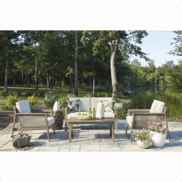 The Barn Cove Grouping 4-piece patio set, featuring a sofa, two chairs, and a coffee table, is arranged on a stone patio overlooking a pond and trees under a clear sky.