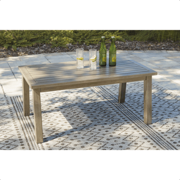 The Barn Cove Grouping 4-piece rectangular wooden outdoor table with four legs stands on a patterned rug, topped with three glass bottles and two glasses with lemon slices. Green foliage and stone pavers are visible in the background.
