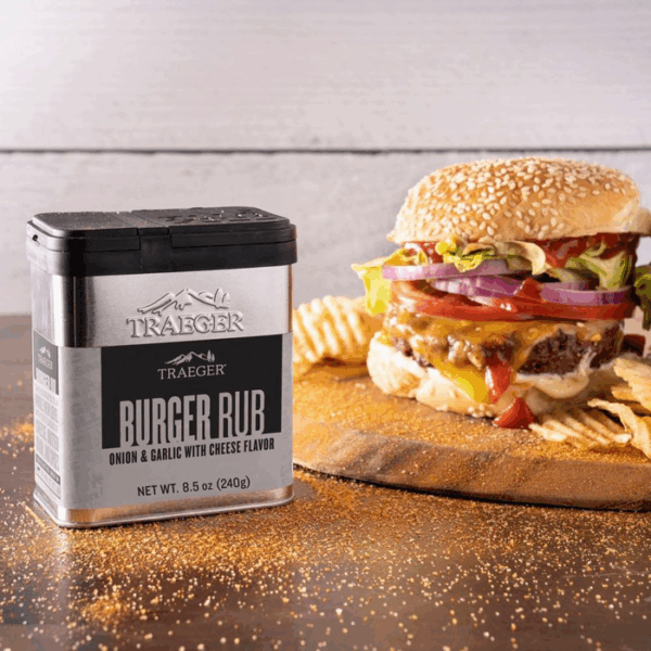 A tin of Traeger SMASH BURGER SEASONING sits on a table sprinkled with seasoning, next to a cheeseburger topped with lettuce, tomato, onion, and served with potato chips on a wooden board.