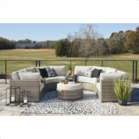 A modern outdoor patio features the Calworth Grouping 8-piece curved wicker sectional with neutral cushions, a round ottoman, black and white pillows, potted plants, lanterns, and a leopard print rug. Grassy field and trees in the background.