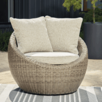 The Danson Large Swivel Lounge, a round wicker outdoor chair with light beige cushions and two large back pillows, is set on a striped rug with plants and a dark wall in the background.