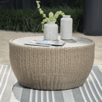 The Danson Cocktail Table, a round wicker outdoor piece with a smooth top, holds two white vases with green foliage and magazines. It sits on a striped rug on the patio, surrounded by lush greenery.