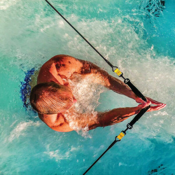A swimmer in blue trunks dives into a pool equipped with the Dual Temp 19 Sierra Shell/Java Cabinet, gripping two resistance bands attached to each side as splashes rise in the clear blue water.