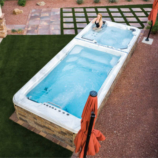 A woman relaxes in the hot tub of the Dual Temp 19 Sierra Shell/Java Cabinet outdoor swim spa, surrounded by grass, stone pavers, and red patio umbrellas.