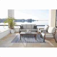 The Emmeline Grouping 4-piece patio set features a cushioned sofa, two armchairs, and a coffee table on a patterned rug. A potted plant sits to the left, overlooking a scenic lake with trees and houses in the background.