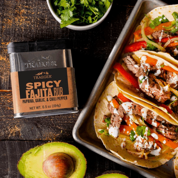 A tin of Traeger FAJITA SEASONING sits beside a tray of steak fajitas loaded with vegetables, cilantro, and sauce. Fresh cilantro and a halved avocado are also on the wooden table.