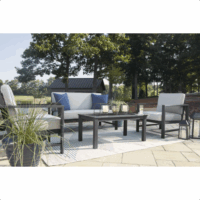 The Fynnegan Grouping 4-piece outdoor patio set features two armchairs, a loveseat, and a rectangular coffee table on a striped rug. Gray cushions and blue accent pillows complete the look; trees and a small shed appear in the background.