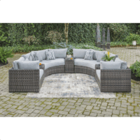 The Harbor Court Grouping 7-piece features a curved gray wicker outdoor sectional with light blue cushions and accent pillows, set around a patterned rug on a stone patio with lush greenery and decorative sculptures in the background.