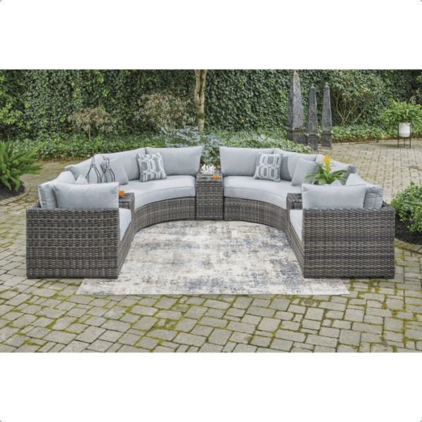 The Harbor Court Grouping 7-piece features a curved gray wicker outdoor sectional with light blue cushions and accent pillows, set around a patterned rug on a stone patio with lush greenery and decorative sculptures in the background.