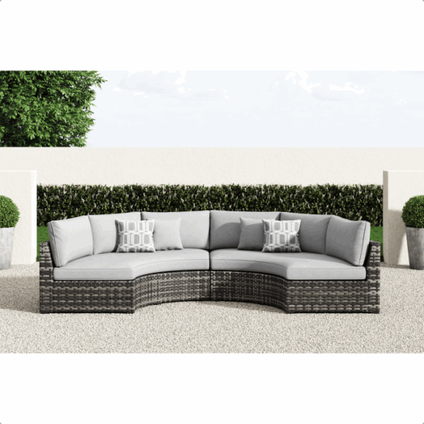 The Harbor Court Grouping 7-piece features a curved outdoor sectional with gray cushions and four patterned throw pillows, displayed on a gravel patio with large potted plants and a green hedge in the background.