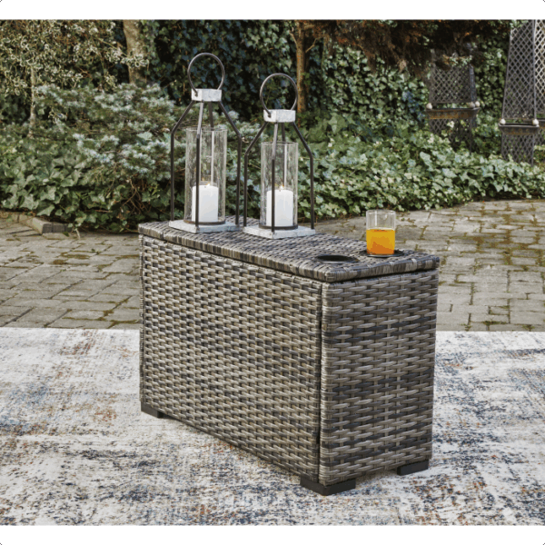 The Harbor Court Grouping 7-piece features a small rectangular wicker storage box on an outdoor rug, topped with glass lanterns holding white candles and a glass of orange juice, set against lush greenery and a stone patio backdrop.